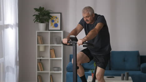 Un-Hombre-De-Mediana-Edad-Está-Entrenando-Con-Una-Bicicleta-Estática-En-Casa.-Plano-Medio-De-Una-Persona-De-Pelo-Gris-Con-Bigote-Y-Ejercicio-Cardiovascular.
